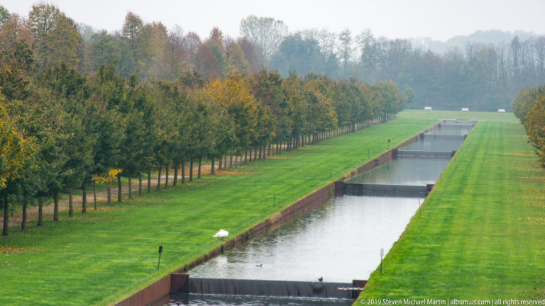 Long rectangular garden canal with stepped drops, flanked by lawns and parallel rows of autumn trees.