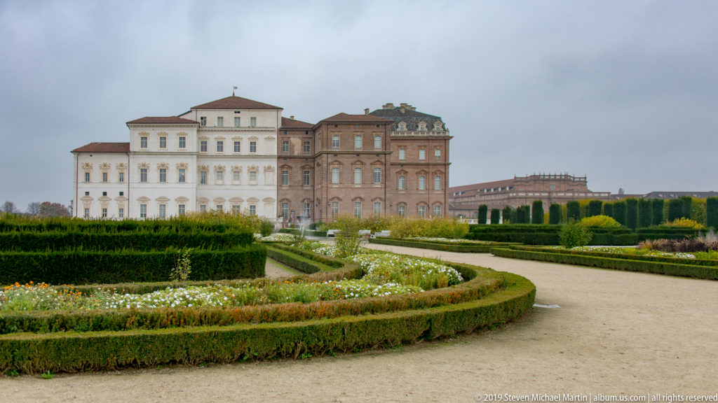 Curving formal parterre with clipped hedges and flowers in front of the pale and brick elevations of La Venaria Reale.