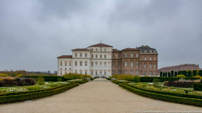 Straight gravel garden path bordered by clipped flower beds leading toward the pale and brick palace façades.