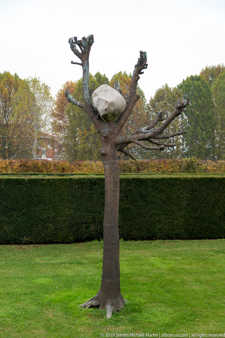 Tree-shaped contemporary sculpture with bare branching arms and a pale suspended form standing on a manicured lawn.