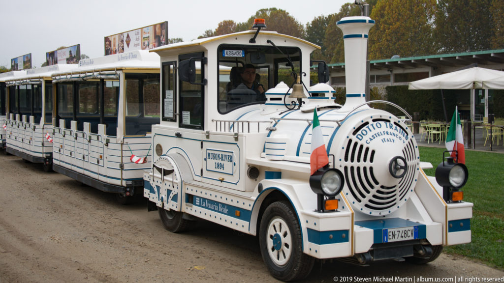 White and blue tourist road train with enclosed passenger cars parked beside the grounds of La Venaria Reale.
