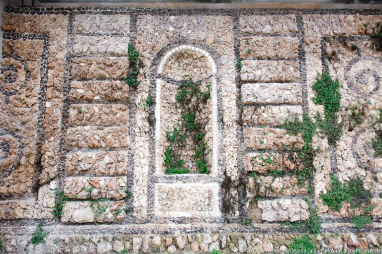 Small arched niche with pale decorative border set into a rough stone wall, with green plants growing in the recess and joints.