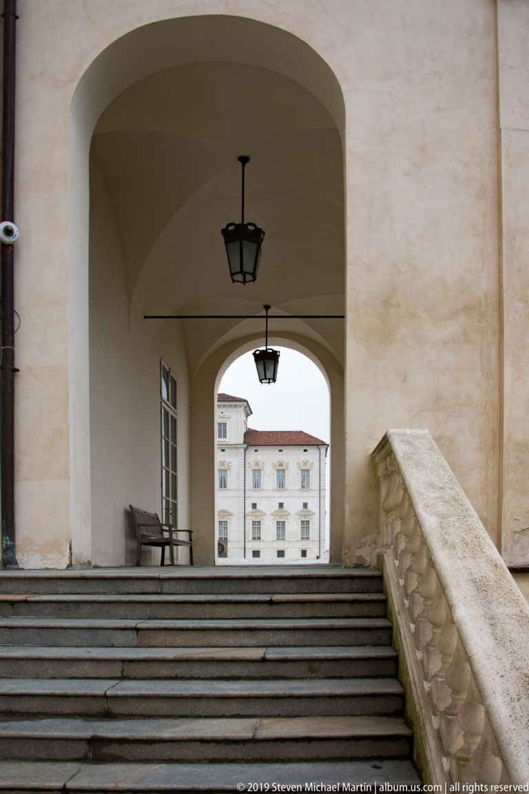 Stone steps beneath a large plastered archway with hanging lanterns and bench, framing a pale palace building beyond.
