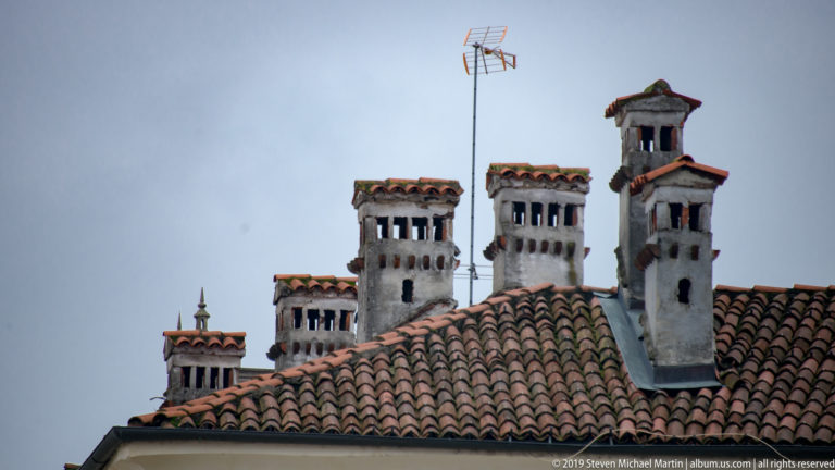 Several tall masonry chimneys with red-tile caps rise above a terracotta roof, with a television antenna against the gray sky.