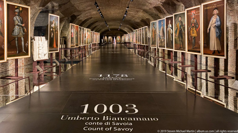 Long vaulted gallery lined with dynastic portraits; floor text reads “1003 Umberto Biancamano, conte di Savoia / Count of Savoy.”