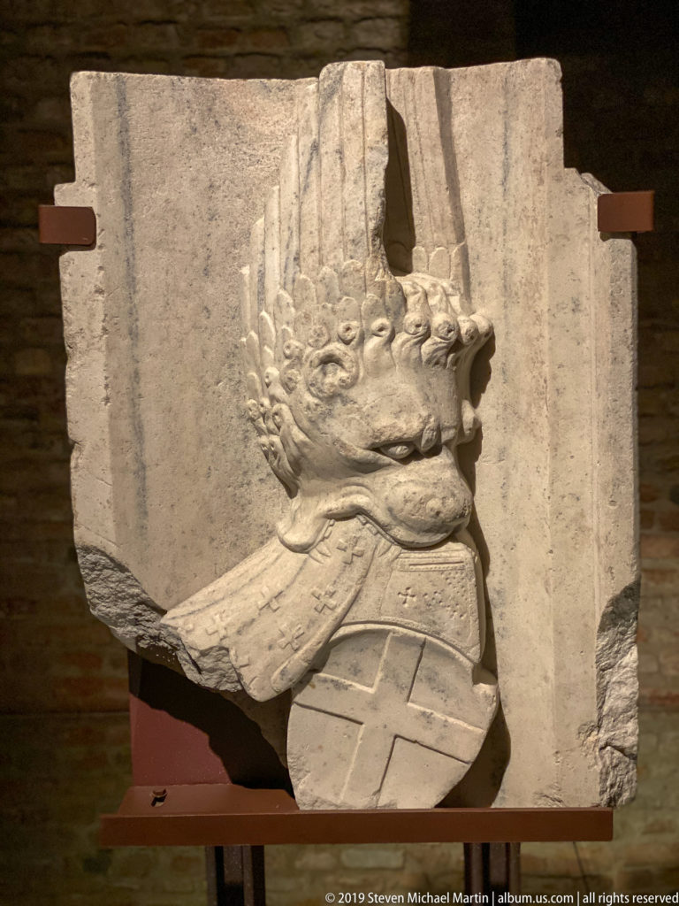 Broken stone relief on a museum mount showing a profile head with a tall crested helmet and part of the upper torso.