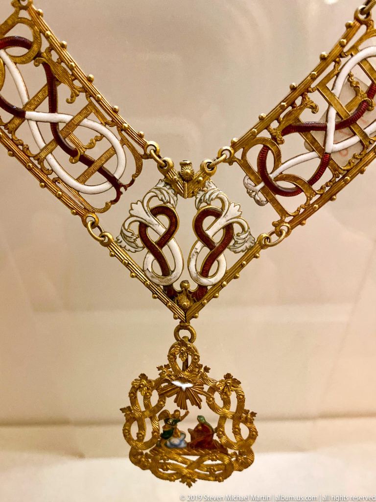 Gold-and-white ceremonial collar with red scroll details and an ornate pendant displayed flat in a museum case.