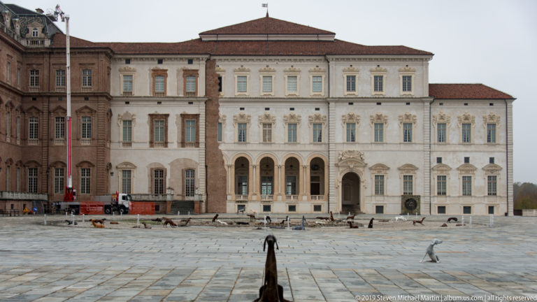 Grand La Venaria Reale courtyard façade with ornate pale central section, brick wings, and animal sculptures on the paving.