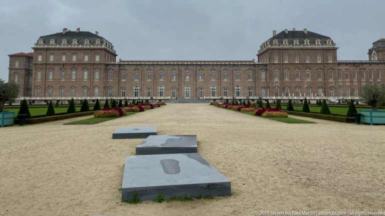 Long symmetrical palace façade beyond a gravel garden axis, with clipped conifers, flower beds, and projecting roofed pavilions.