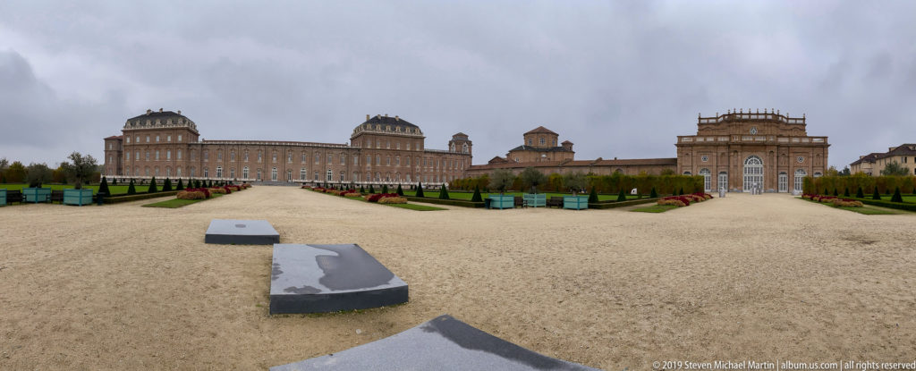 Panoramic garden view toward La Venaria Reale, with long brick wings, towers, chapel, gravel paths, and low foreground markers.