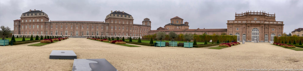 Wide view across gravel paths and formal plantings toward the brick palace and chapel buildings of La Venaria Reale.