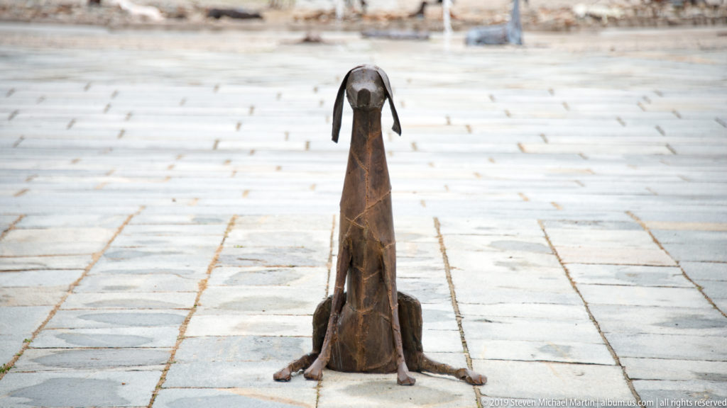 Stylized seated metal dog sculpture with long neck and drooping ears on pale courtyard paving.