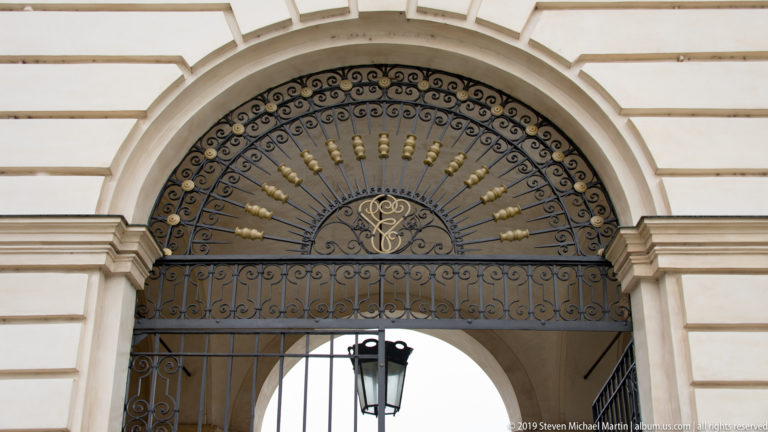 Semicircular wrought-iron gate fan with radiating scrollwork, gilded ornaments, central monogram, and hanging lantern.