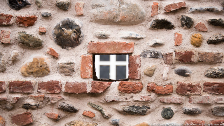 Small white cross-shaped opening framed by red brick in a rough wall of fieldstone, brick fragments, and mortar.