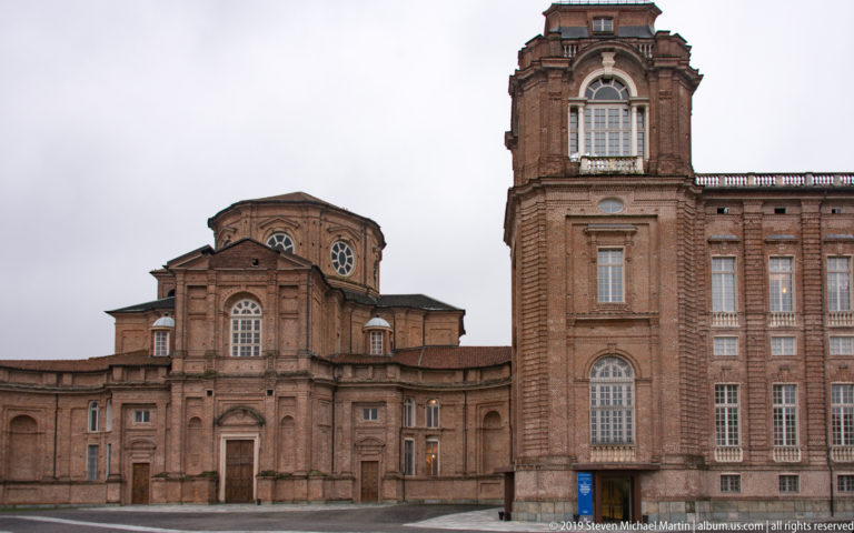 Brick façade of the Church of Sant'Uberto beside an adjoining La Venaria Reale palace wing under an overcast sky.
