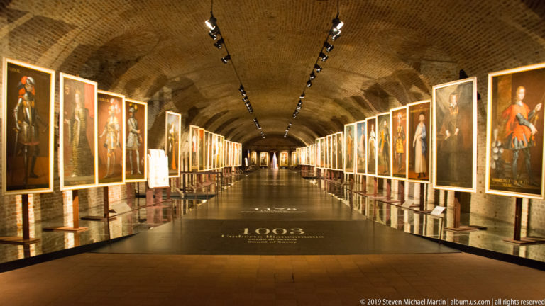 Long brick-vaulted gallery lined with illuminated full-length portraits, with the date 1003 marked on the floor.