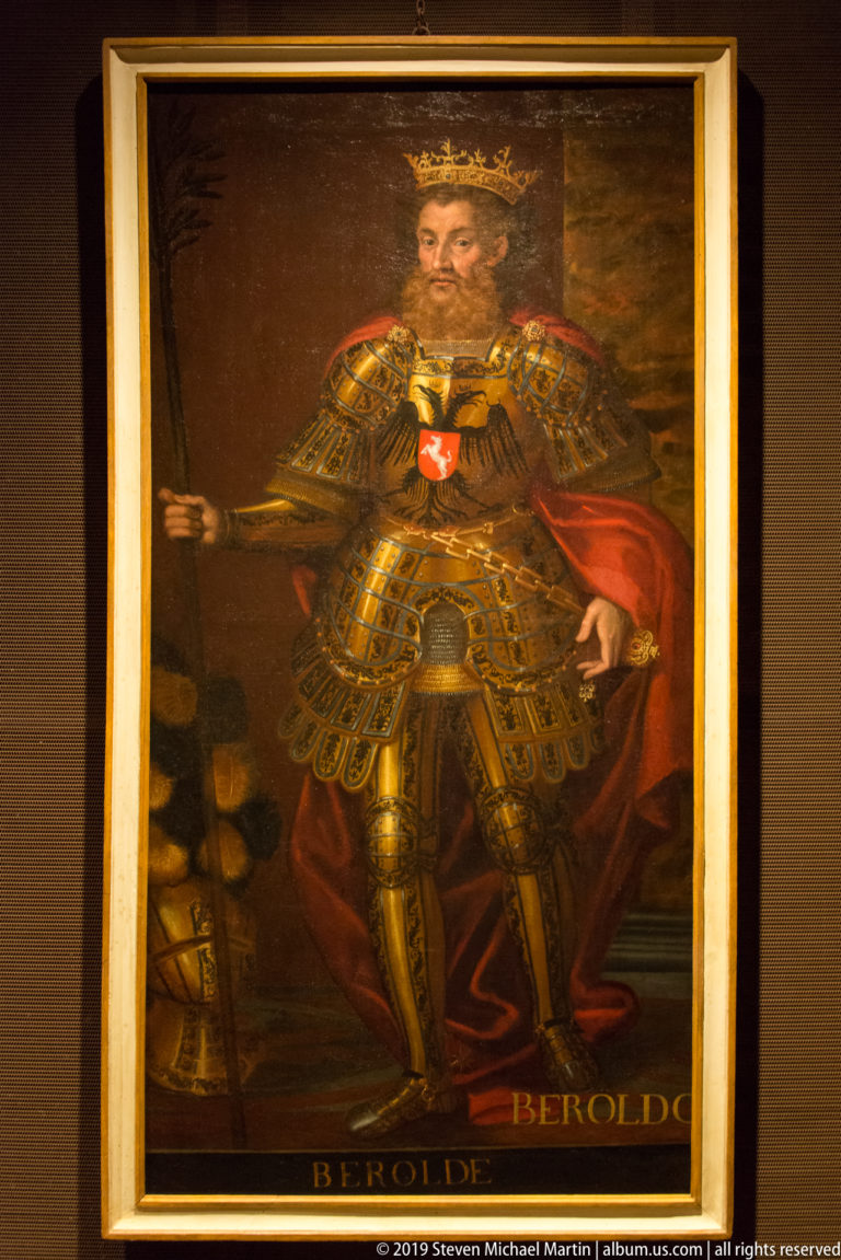 Full-length painted portrait labeled Beroldo, wearing elaborate armor and red cloak and holding a long staff.