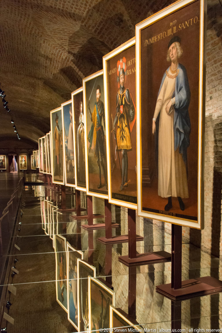 Oblique row of illuminated full-length Savoy portraits on stands inside a dark brick-vaulted gallery.