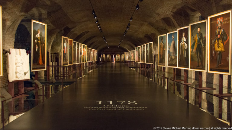 Long brick-vaulted gallery lined with illuminated full-length Savoy portraits and a floor date of 1178.