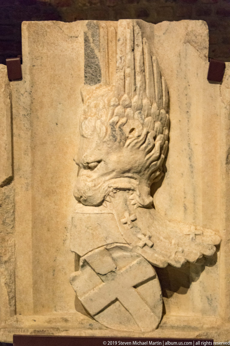 Weathered stone relief of a crested bird or griffin-like heraldic creature above a shield-shaped form.