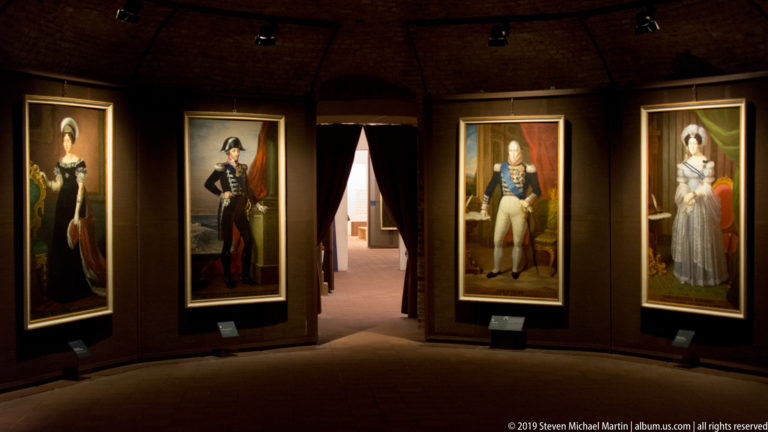 Four illuminated full-length historical portraits displayed along a dark curved gallery wall.