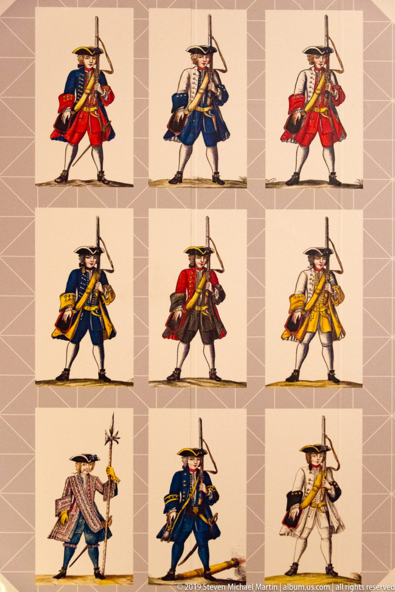 Nine illustrated historical soldiers in varied red, blue, yellow, and white uniforms carrying long weapons.