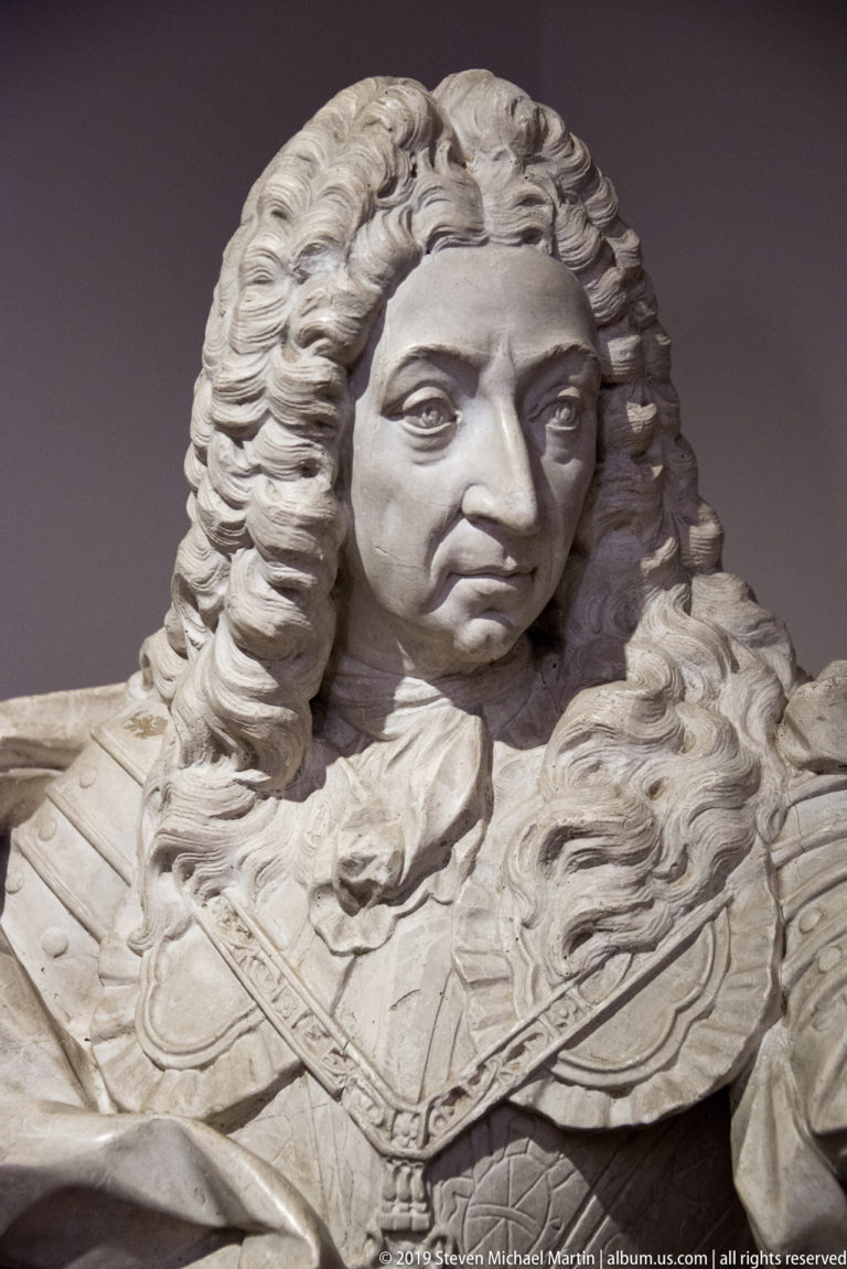 Close view of a marble nobleman’s bust with long curled hair, broad collar, armor, and a severe expression.