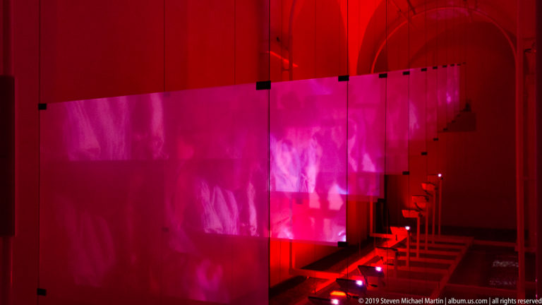 Red and magenta translucent screens with projected human silhouettes receding through a dark vaulted interior.