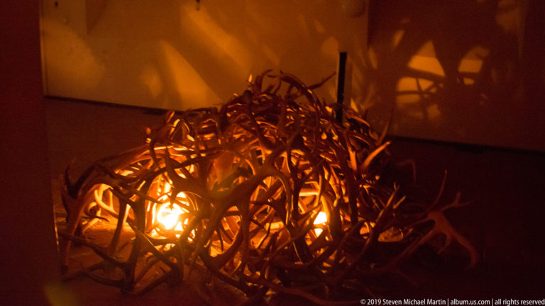 Glowing nest-like sculpture of tangled branches casting large amber shadows on surrounding walls.