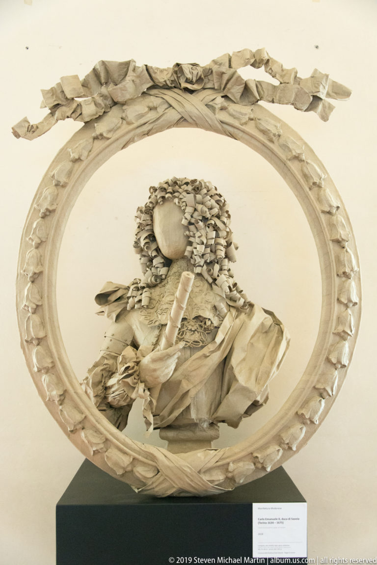 White sculpted bust of a courtly woman with elaborate curls inside an oval wreath crowned by a bow.