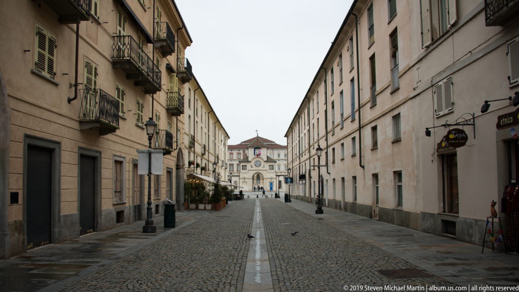 Long cobblestone street with balconies, shops, and lamps leading toward the distant clock gate of La Venaria Reale.
