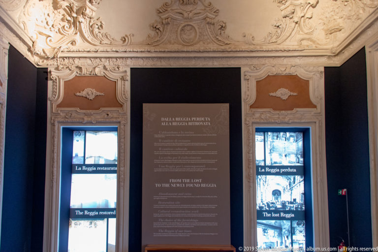 Interpretive panel about the lost Reggia between two illuminated displays, beneath ornate stucco decoration.