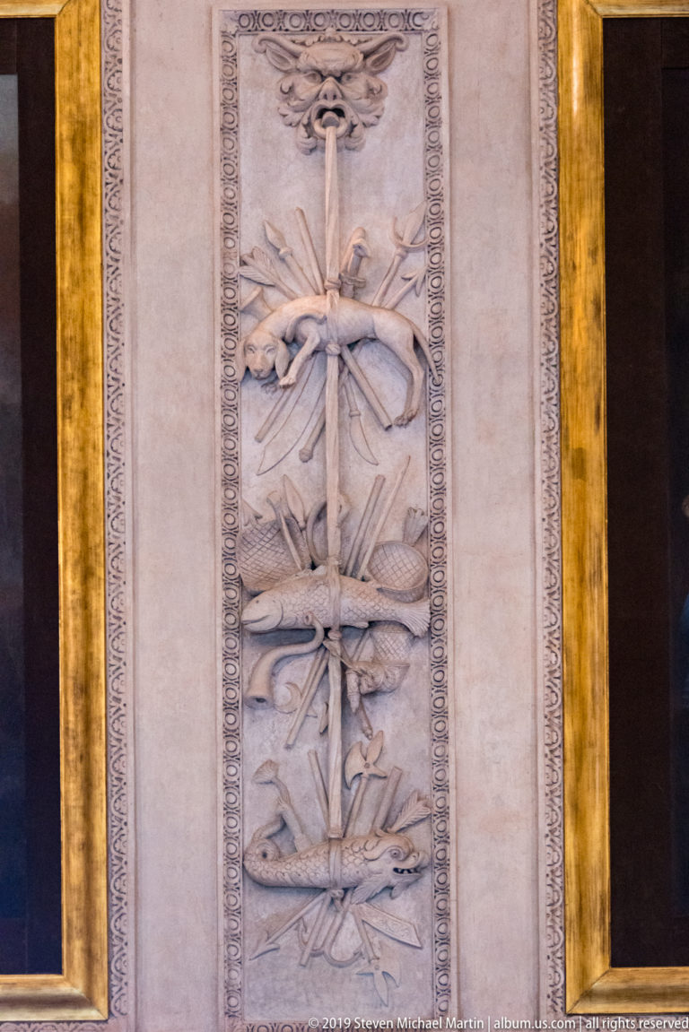 Vertical pale stucco relief of crossed weapons, standards, and military equipment within decorative borders.