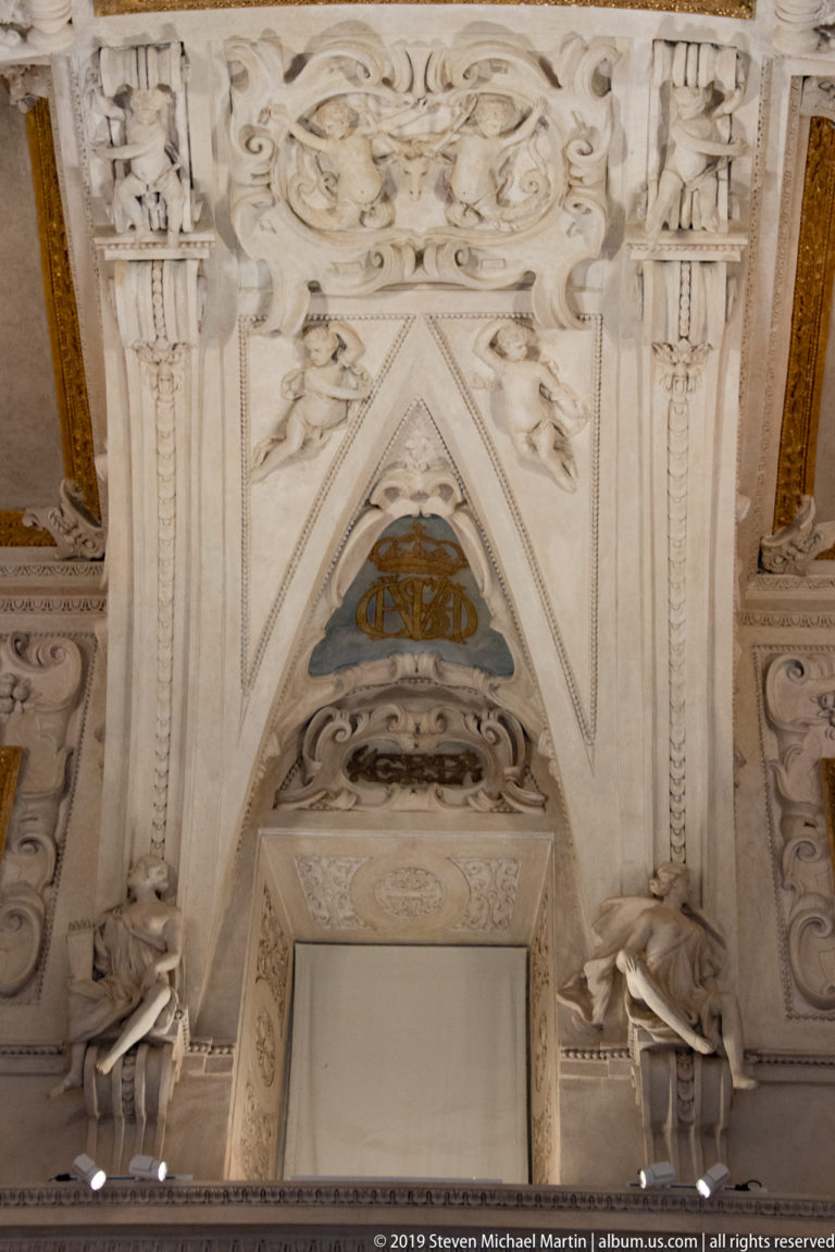 Ornate pale stucco overdoor with central heraldic cartouche and a small painted triangular panel above a doorway.