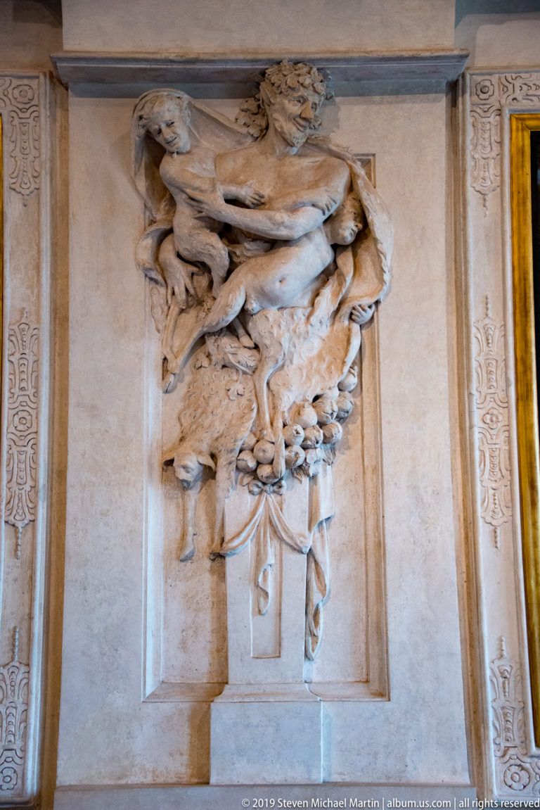 Muscular male stucco figure in high relief above clustered decorative objects and foliage.