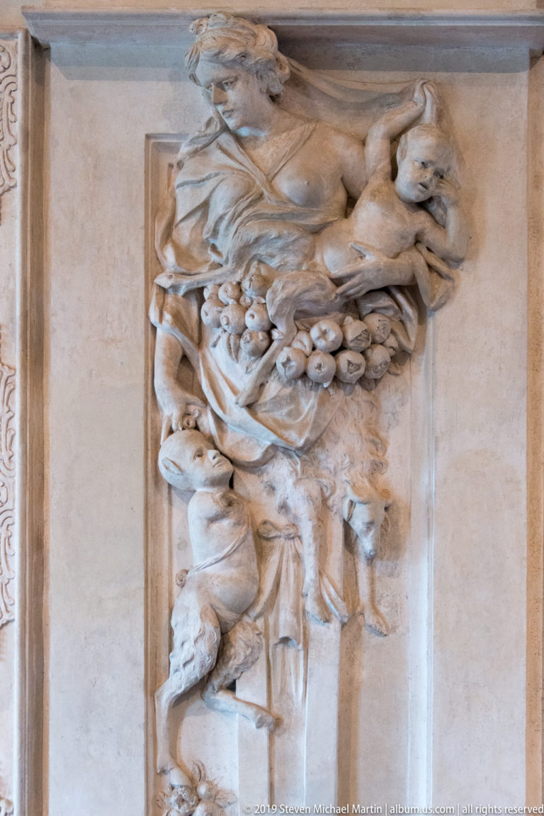 Stucco female figure above a dense cluster of fruit, flowers, and drapery on a palace wall.
