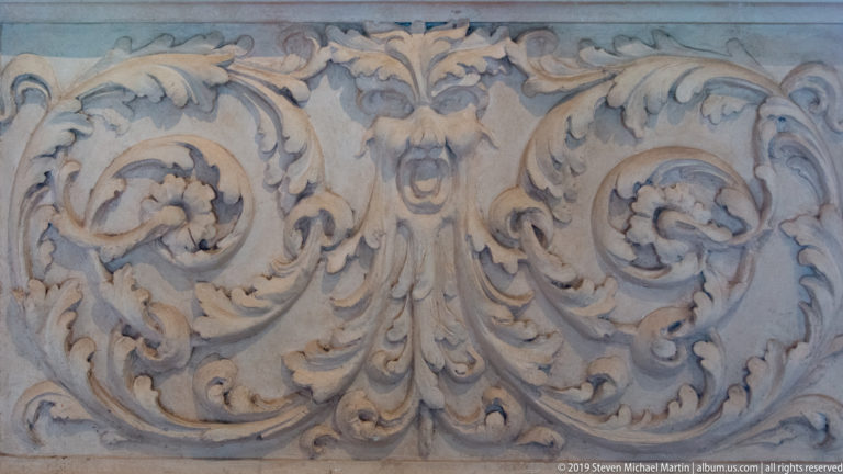 Close detail of symmetrical acanthus scrolls in pale stucco centered on a small grotesque mask.