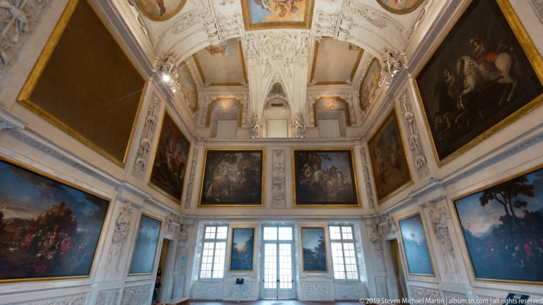 High palace gallery with large framed paintings hung around tall windows beneath an ornate white ceiling.