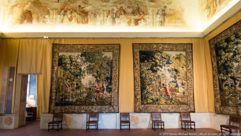 Palace room with three large wooded-scene tapestries hanging above a row of simple chairs.