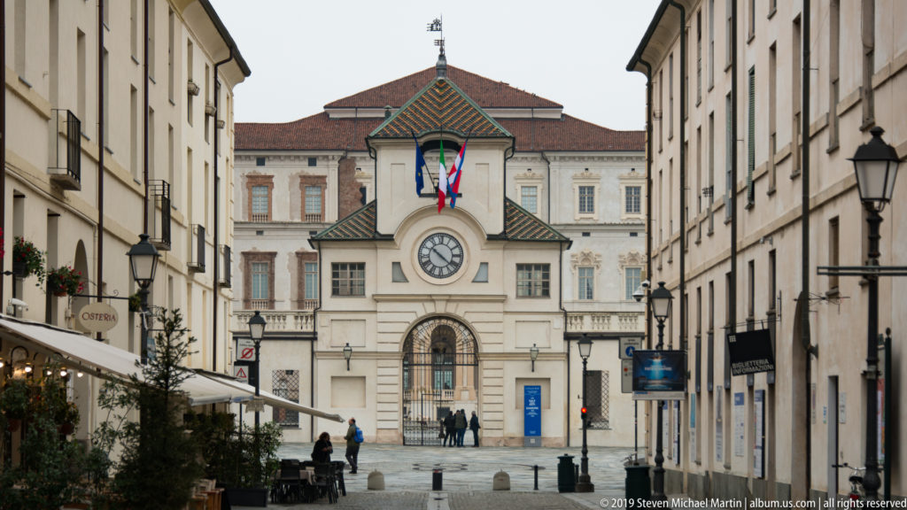 Historic town street lined with shops and lamps leading toward La Venaria Reale’s clock gate.