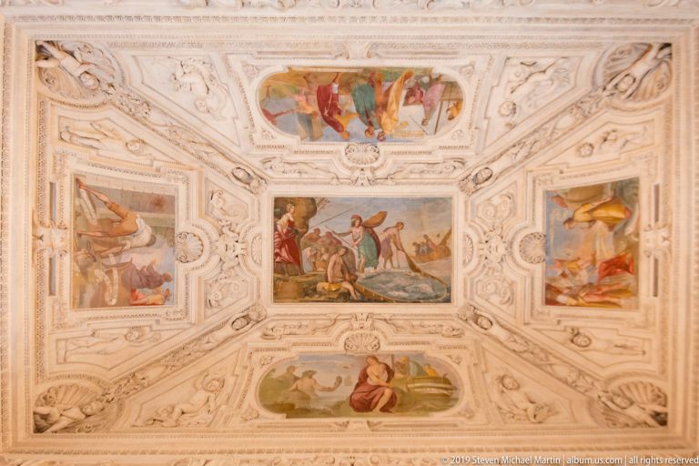 Ceiling with a central rectangular fresco and four surrounding figurative panels framed by dense white stucco.