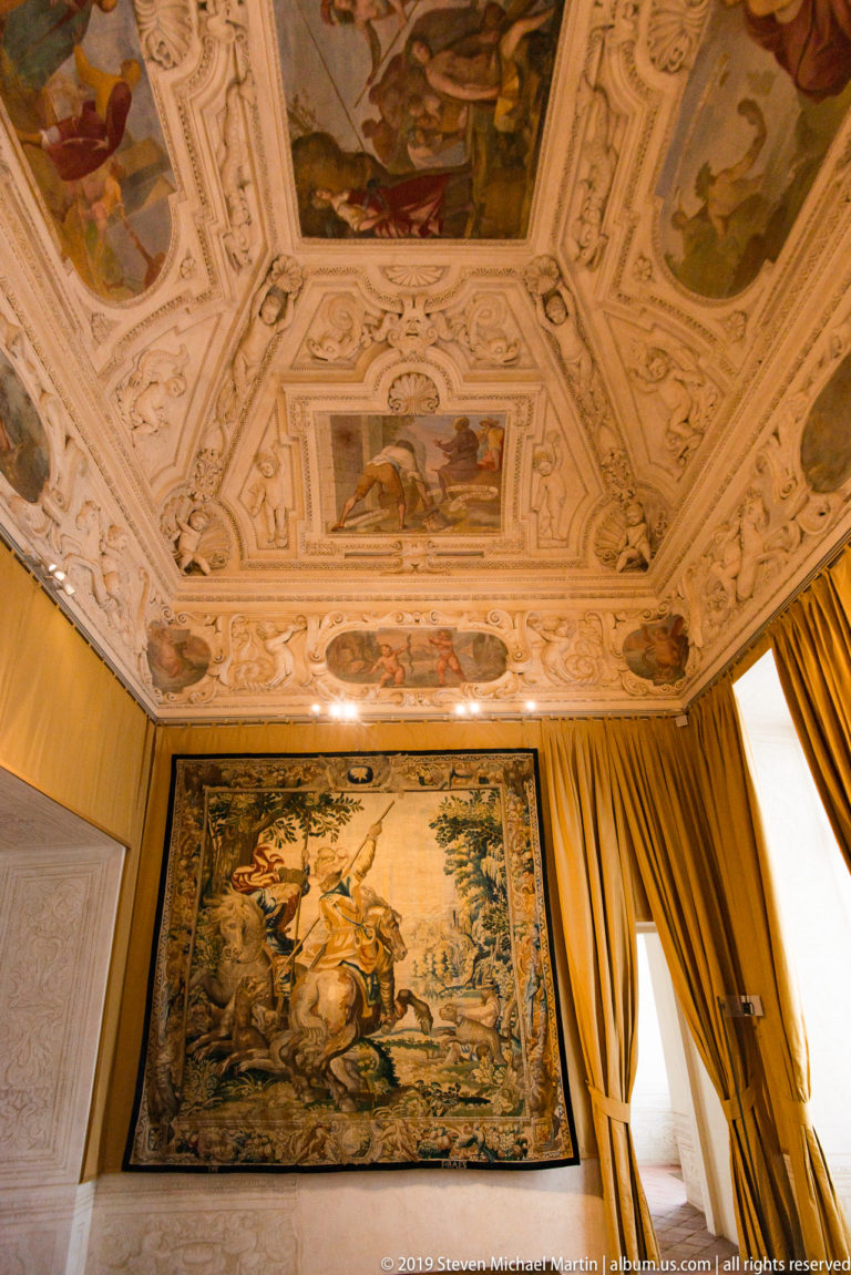 Large hunting tapestry beneath a richly painted and white-stucco ceiling, framed by gold curtains.