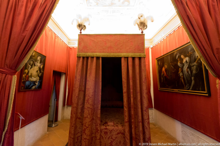 Red-canopied bed in a crimson palace chamber with heavy curtains, framed paintings, and patterned carpet.