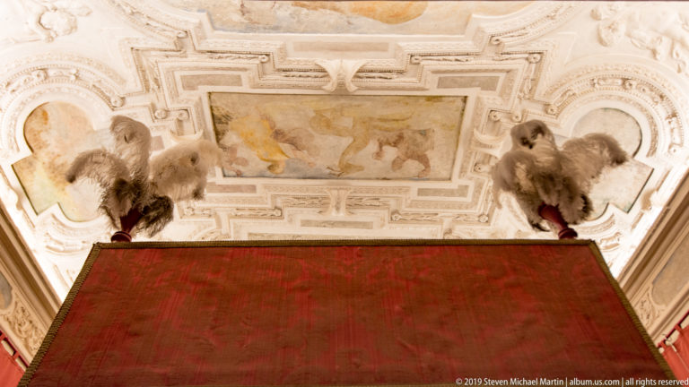 Top of a red bed canopy beneath an ornate frescoed and stuccoed ceiling with sculptural finials at the corners.