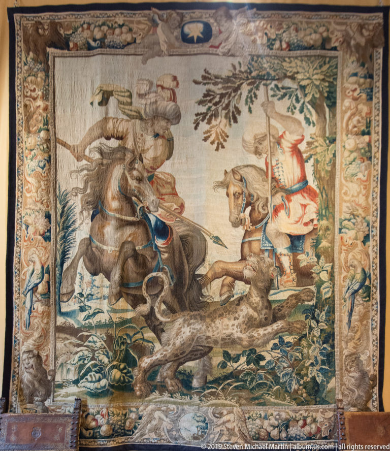 Tapestry with a mounted hunter, running hounds, and another figure in a wooded landscape.