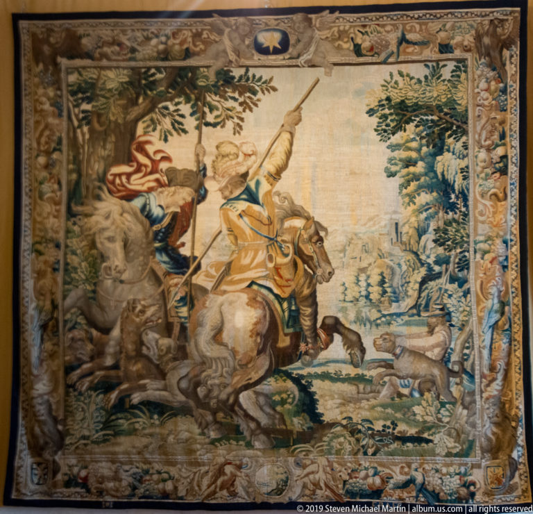 Tapestry of mounted hunters with dogs pursuing game through a wooded landscape.
