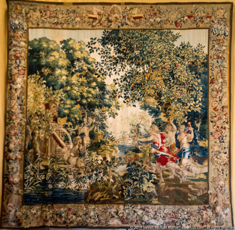 Large tapestry with brightly dressed figures beside water in a dense wooded landscape, surrounded by an ornate border.