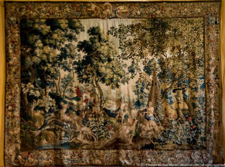 Large tapestry showing a dense wooded landscape with small figures and animals, enclosed by an ornate border.