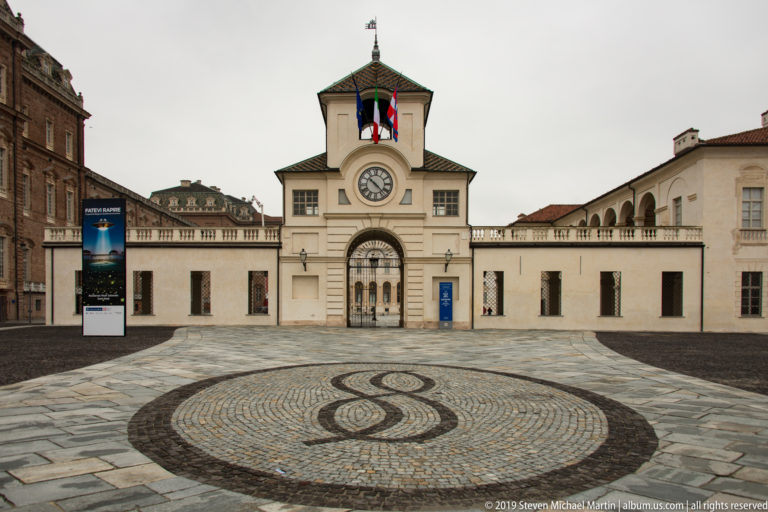 Centered clock gate with flags and arched entry aligned behind a circular looping cobblestone pavement pattern.