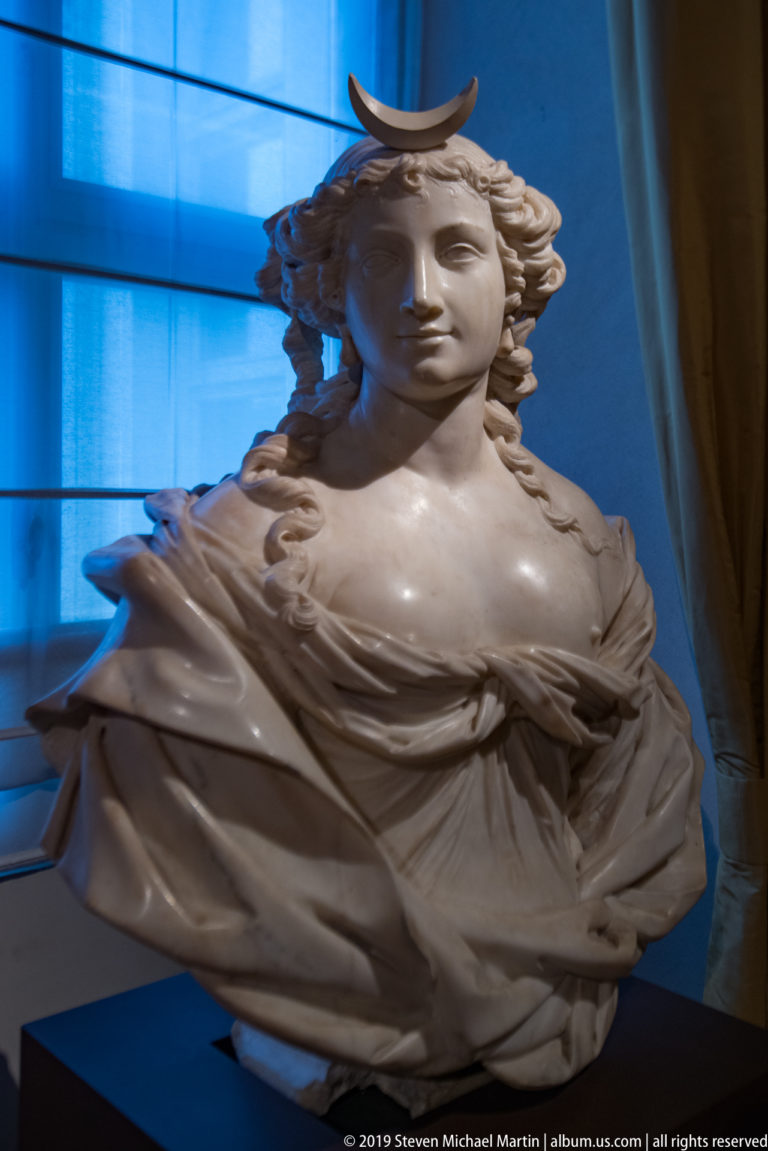 White marble bust of a young woman with flowing hair, drapery, and a crescent-shaped ornament above her forehead.